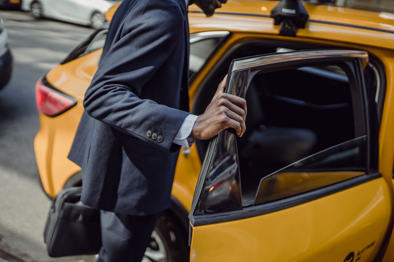 our-experience A businessman in a suit opens the door to a yellow taxi on a city street during the day.