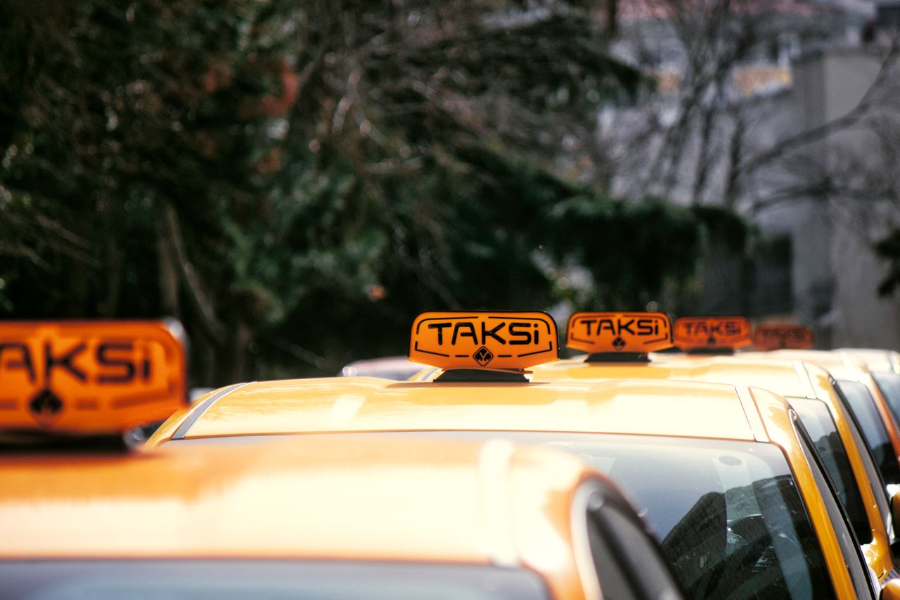services-03 A row of yellow taxis with 'Taksi' signs parked on a city street in daylight.